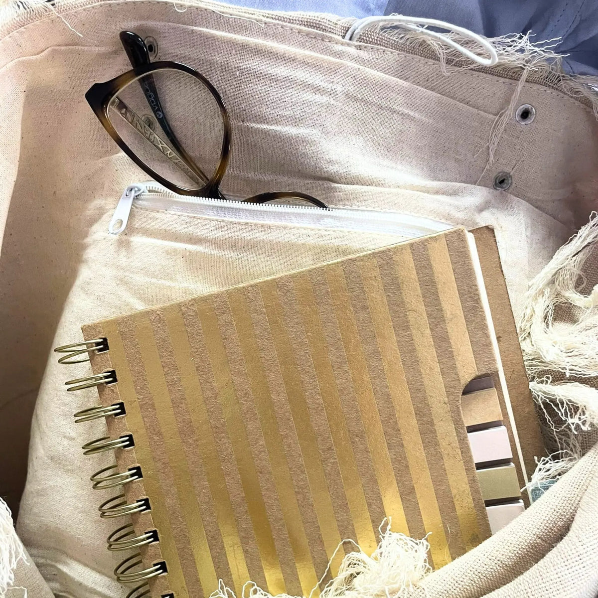 Interior de bolso de tela natural con bordes desflecados, conteniendo una libreta a rayas y gafas de pasta carey.