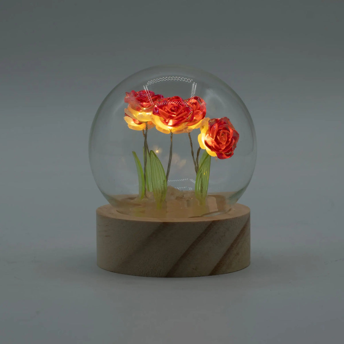 Domo de cristal con cinco rosas rojas iluminadas por luces LED, sobre una base redonda de madera. Estilo decorativo.