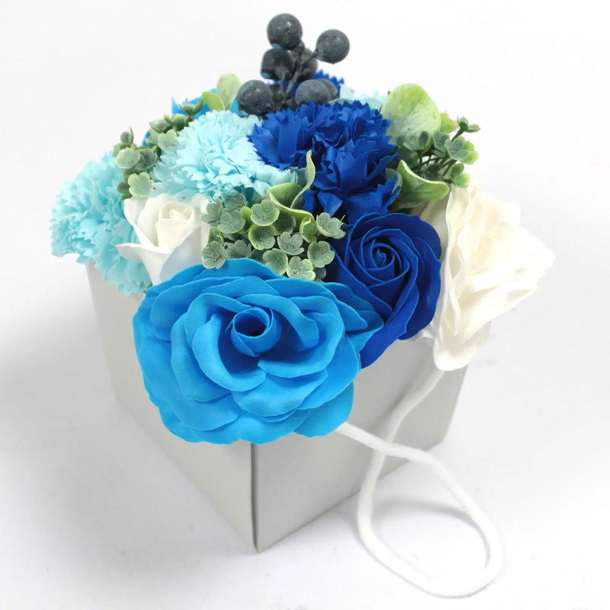 Ramo de flores de jabón con rosas y claveles en tonos azules y blancos, decorado con bayas y hojas, en caja cuadrada.