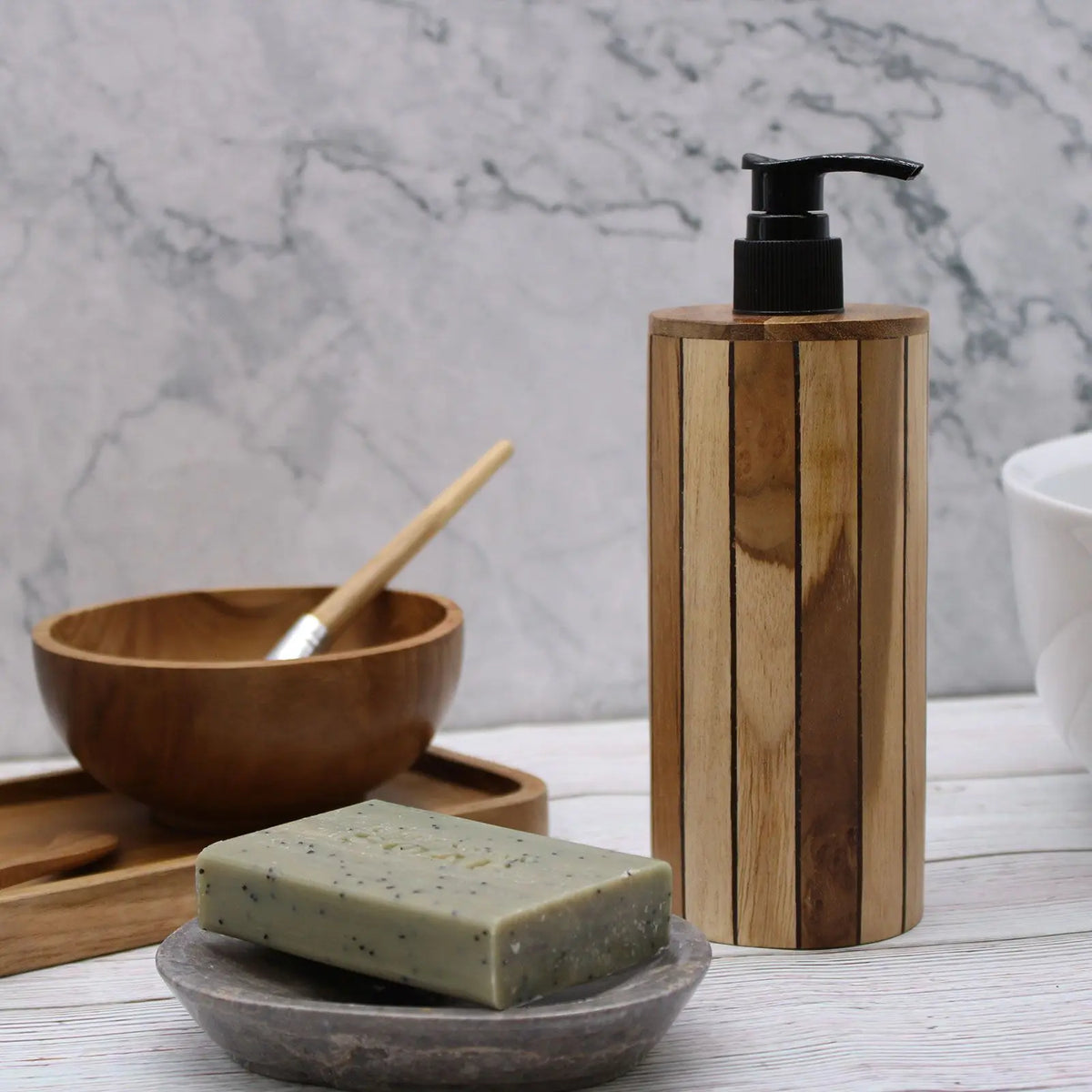 Dispensador de jabón líquido cilíndrico de madera de teca natural con dosificador negro sobre fondo de baño.
