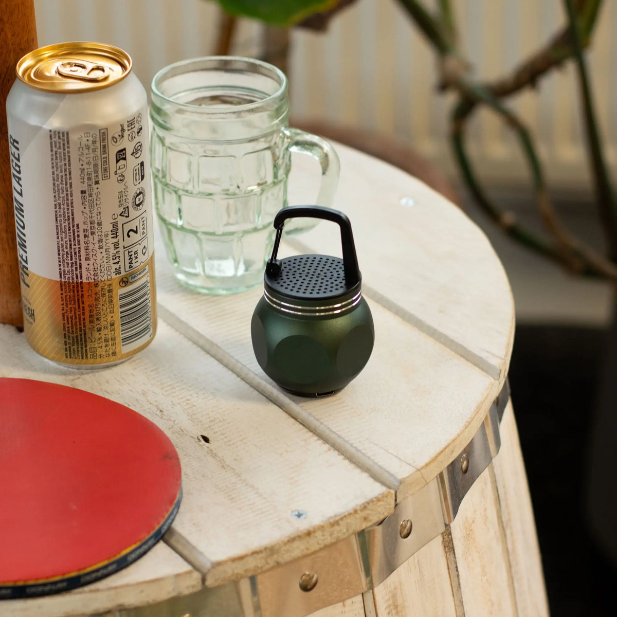 Mini altavoz Bluetooth verde de Ancient Wisdom, metálico, forma redonda con gancho superior sobre mesa de madera.
