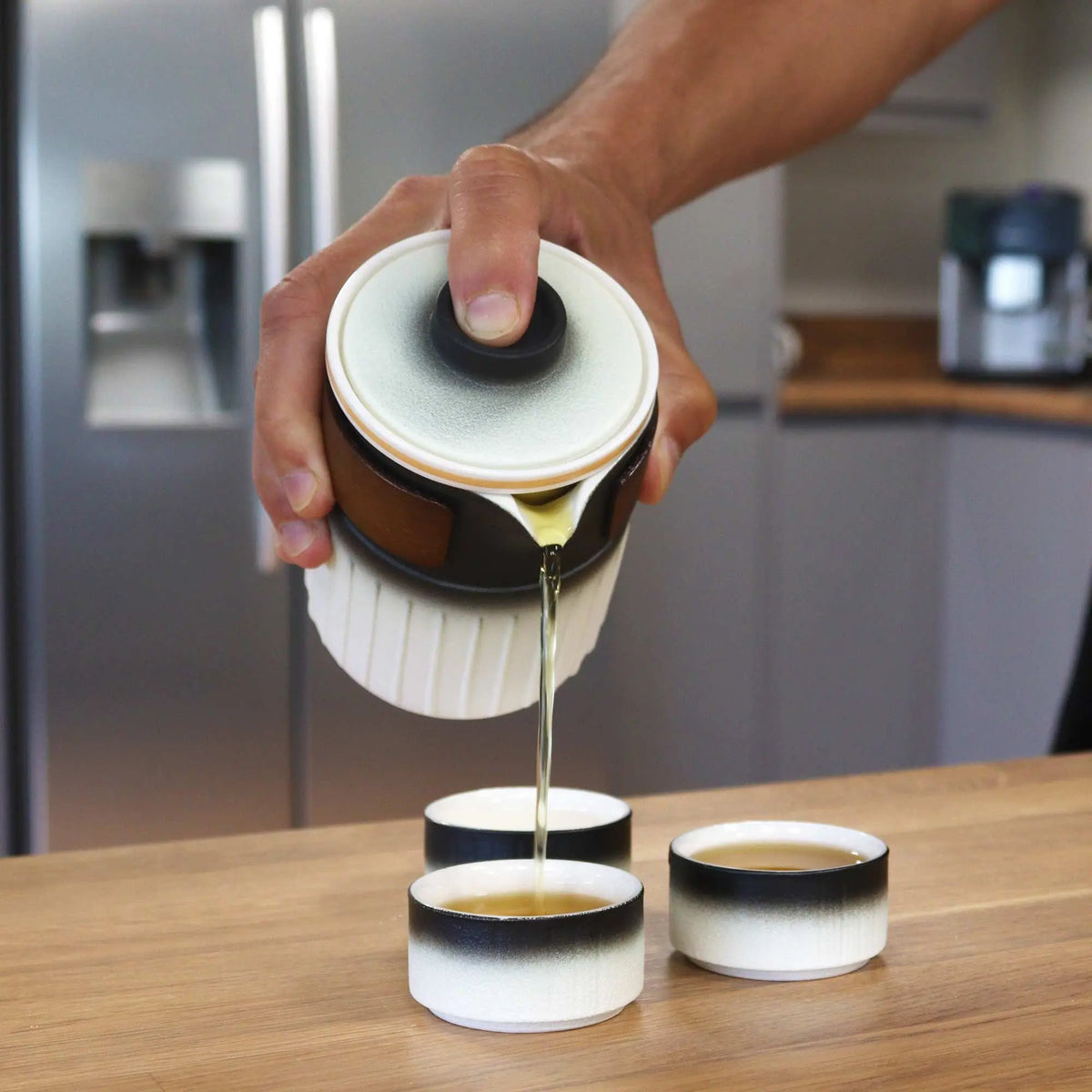 Tetera de cerámica blanca y negra con detalle de cuero sirviendo té en tazas a juego sobre mesa de madera.