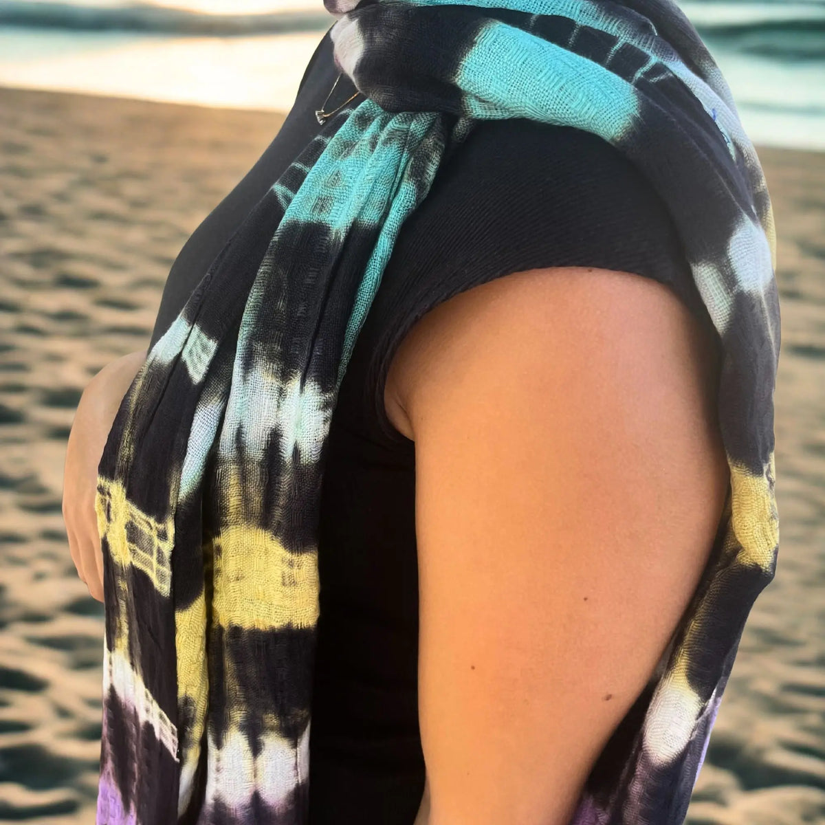 Estola de lino y rayón con rayas de colores, estilo boho, usada sobre los hombros en una playa al atardecer.