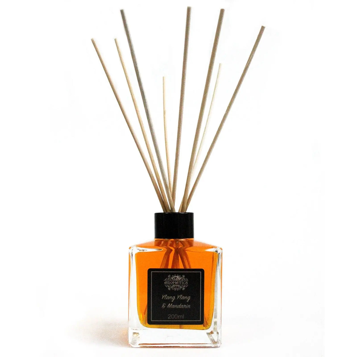 Difusor de varillas con frasco cuadrado de vidrio, líquido ámbar, etiqueta negra y varas de madera sobre fondo blanco.
