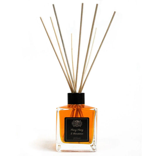 Difusor de varillas con frasco cuadrado de vidrio, líquido ámbar, etiqueta negra y varas de madera sobre fondo blanco.