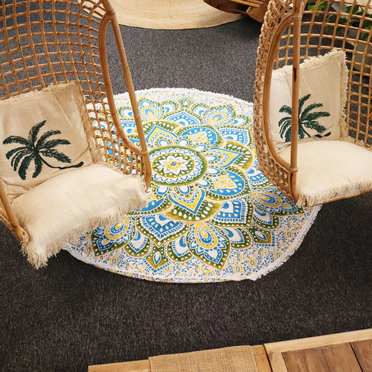 Tapiz de algodón redondo con diseño de mandala en tonos amarillo y azul, sobre suelo gris entre dos sillas colgantes.