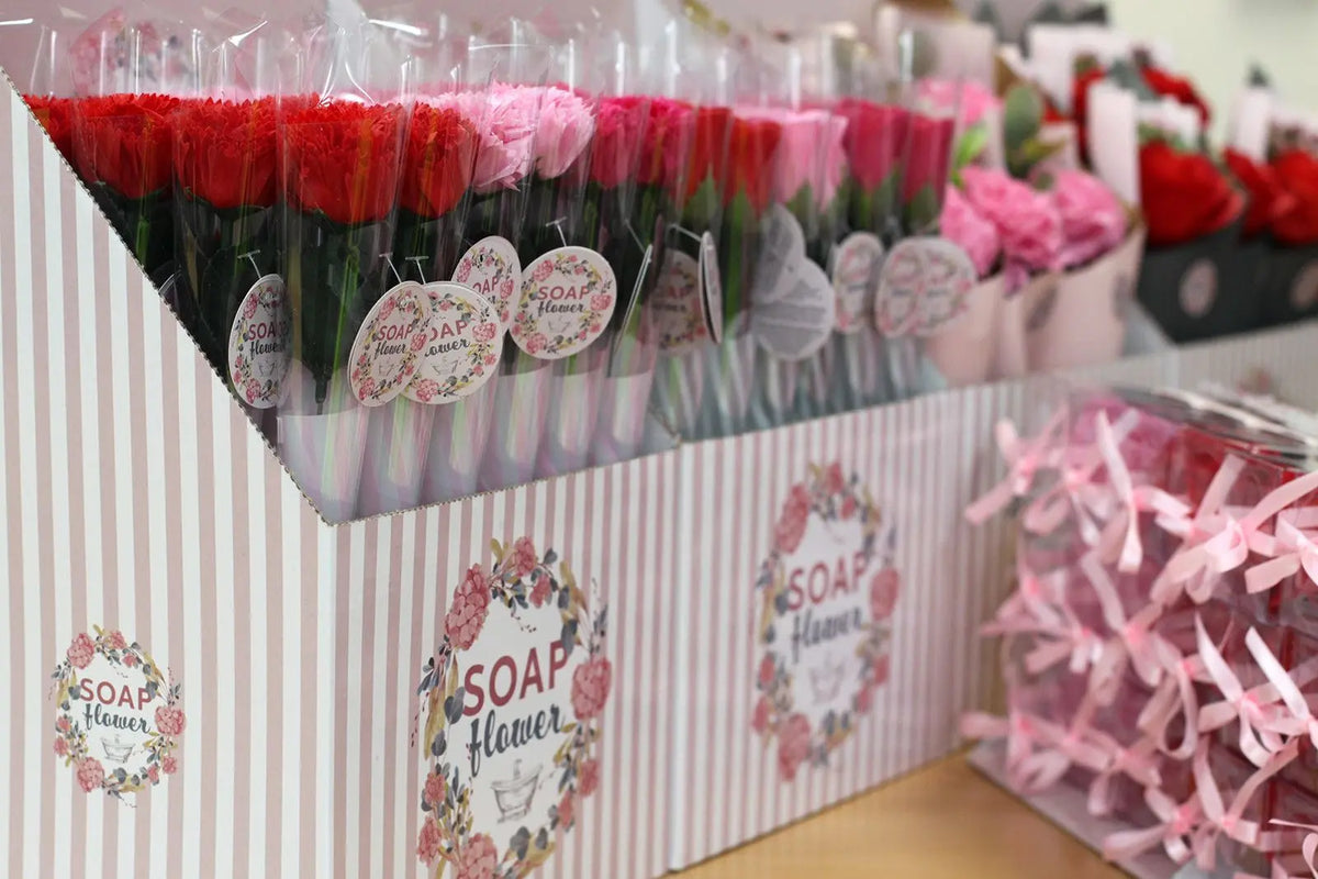 Rosas de jabón envueltas individualmente en tonos rojos y rosas, expuestas en cajas de cartón a rayas para venta.