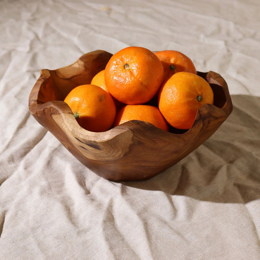 Frutero artesanal de madera de teca con forma orgánica irregular, relleno de mandarinas sobre fondo de tela beige.