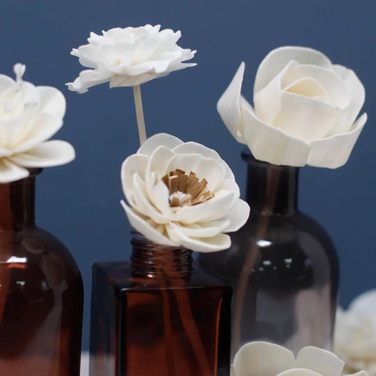 Flores blancas de madera con tallo en frascos de vidrio marrón sobre fondo azul, estilo decorativo.