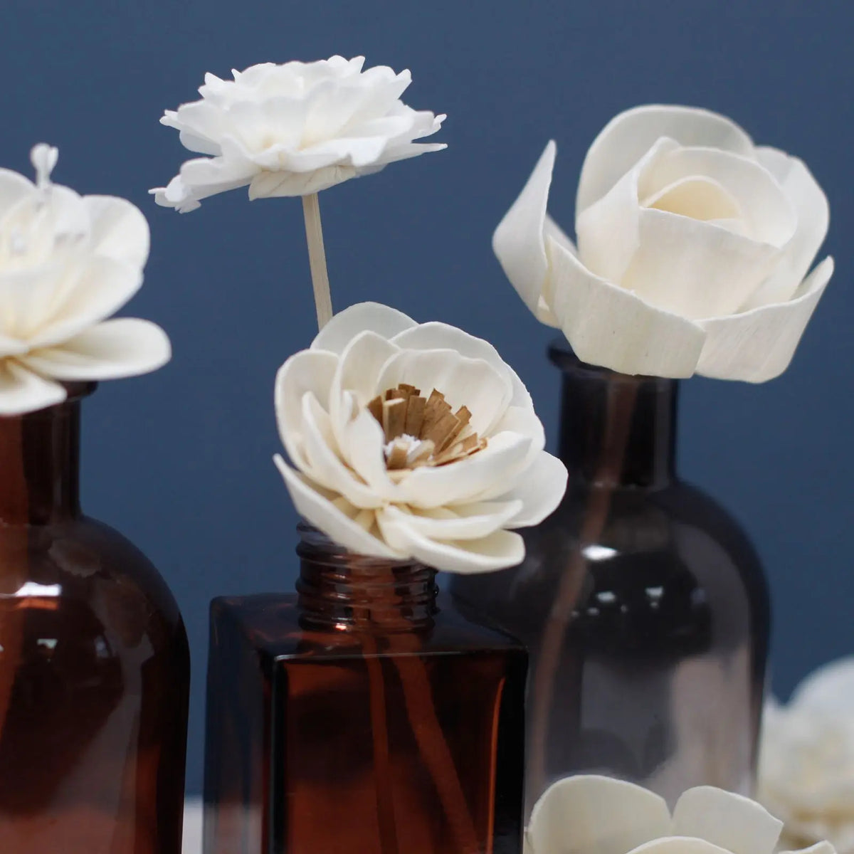 Flores difusoras blancas hechas a mano sobre varillas, presentadas en frascos de vidrio ámbar sobre fondo azul oscuro.