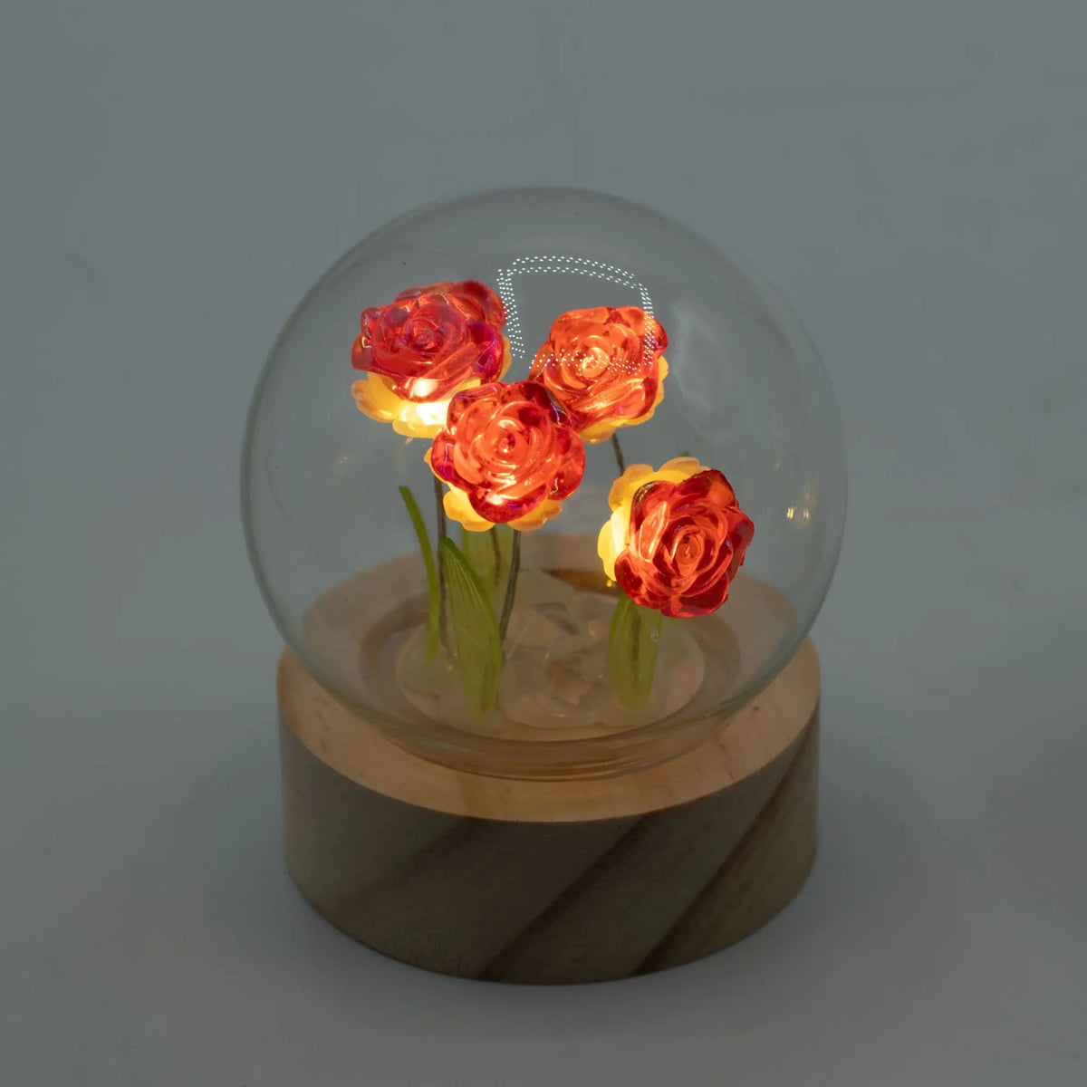 Domo de cristal con 4 rosas rojas LED iluminadas sobre base de madera circular. Estilo elegante y cálido.