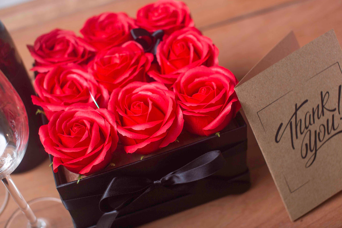 Caja cuadrada negra con 9 rosas rojas de jabón, atada con un lazo y junto a una tarjeta de regalo sobre mesa de madera.
