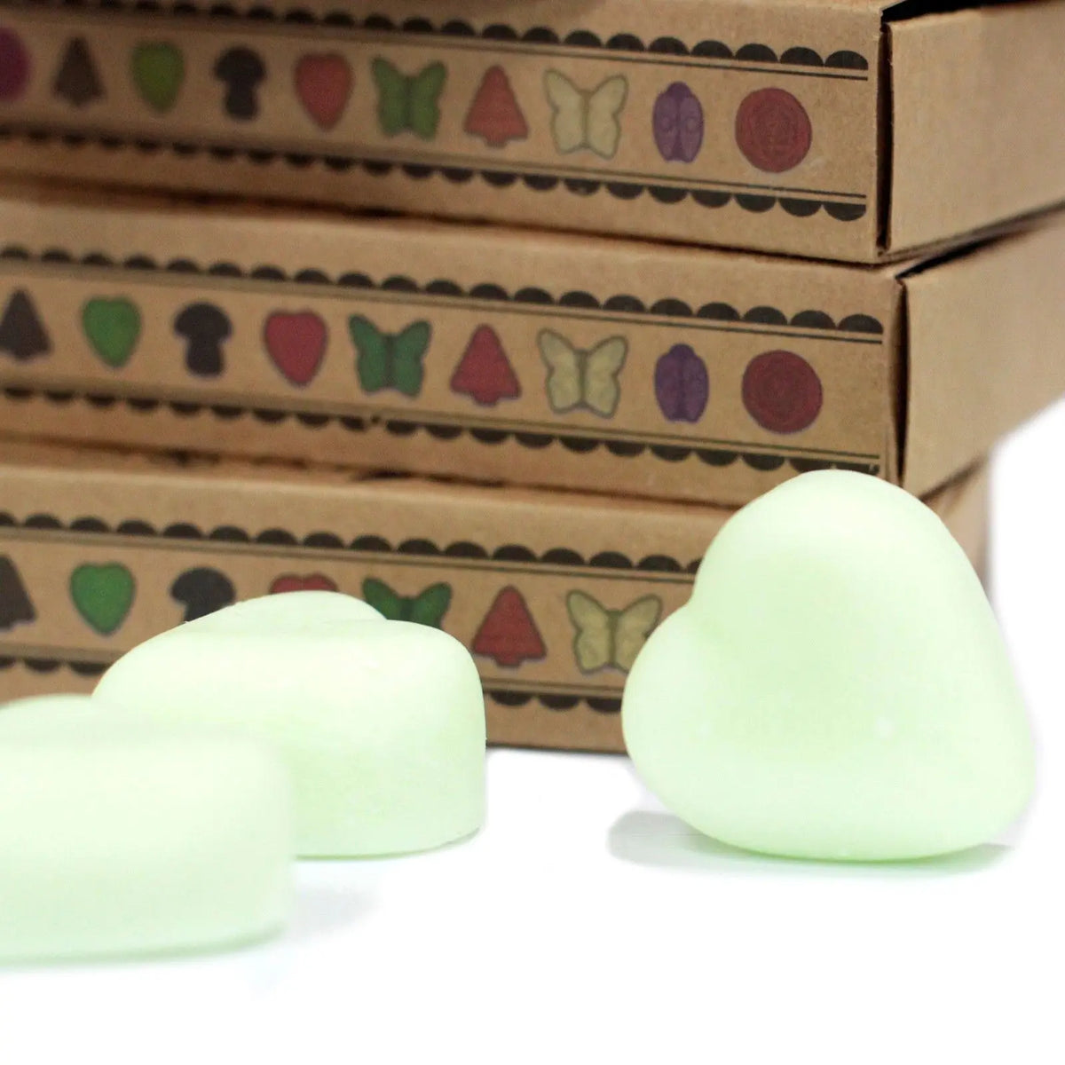 Tres ceras de soja en forma de corazón color verde claro frente a cajas de cartón con dibujos de insectos y frutas.