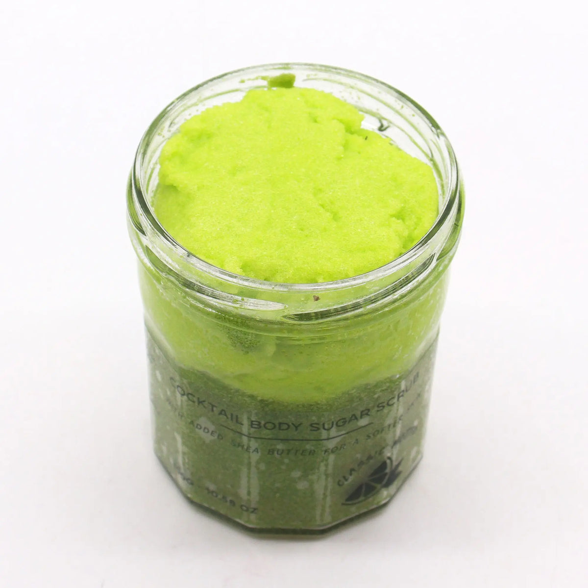 Frasco de vidrio abierto con exfoliante de azúcar color verde brillante, textura granulada sobre fondo blanco.