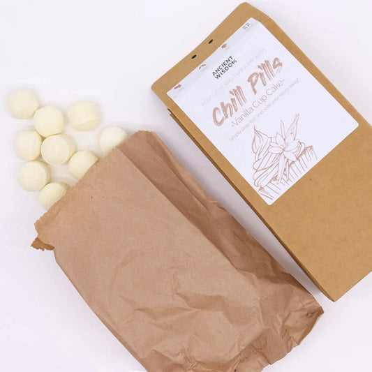 Mini bombas de baño color crema saliendo de una bolsa de papel marrón junto a su caja de regalo sobre fondo blanco.