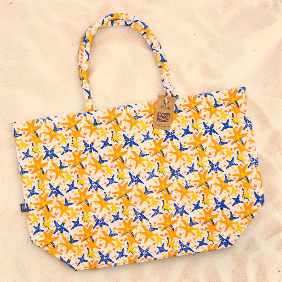 Bolso de tela grande con estampado de estrellas de mar azules y amarillas sobre fondo blanco, apoyado en la arena.