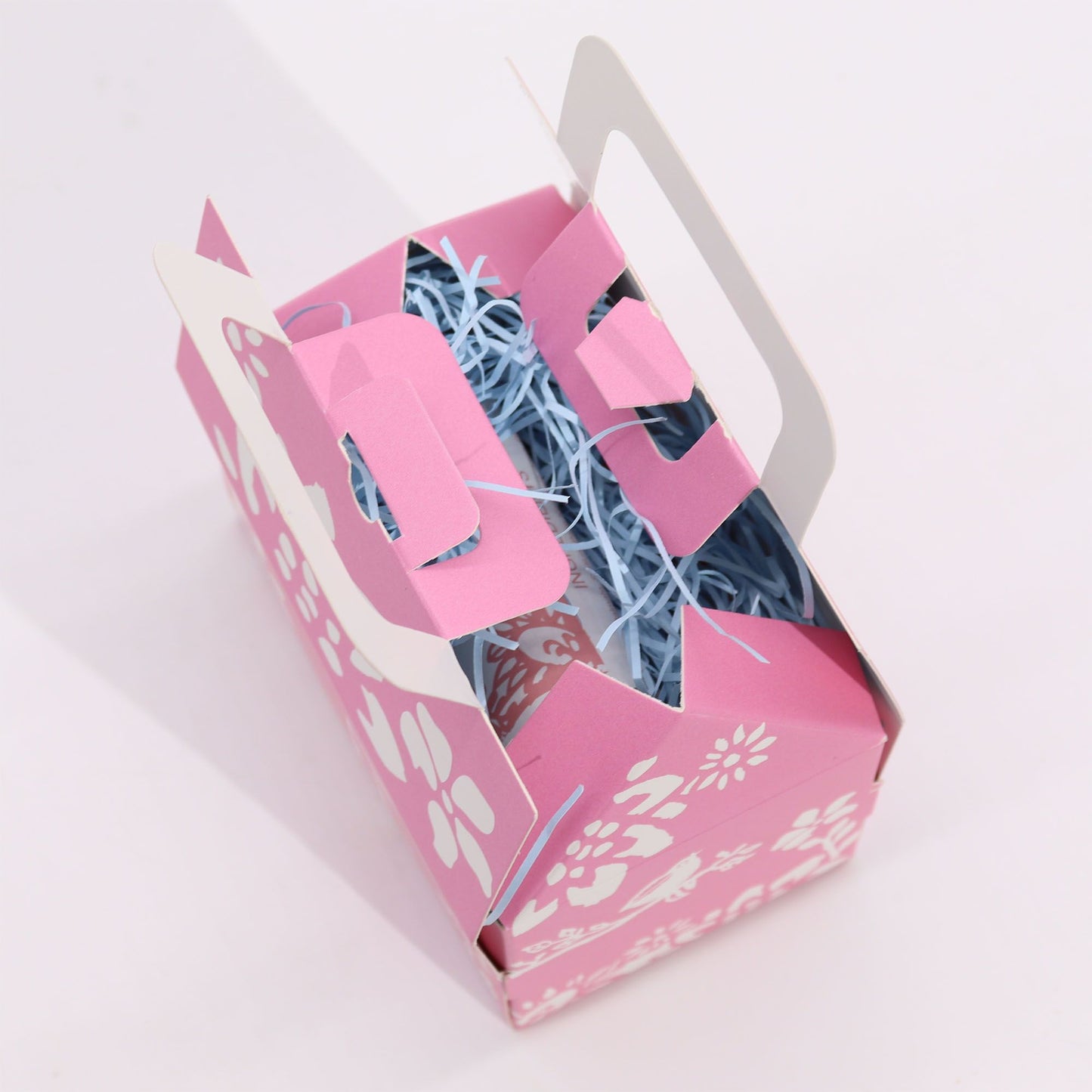 Caja de regalo rosa con flores blancas, asa integrada y relleno de papel azul claro, vista desde arriba.