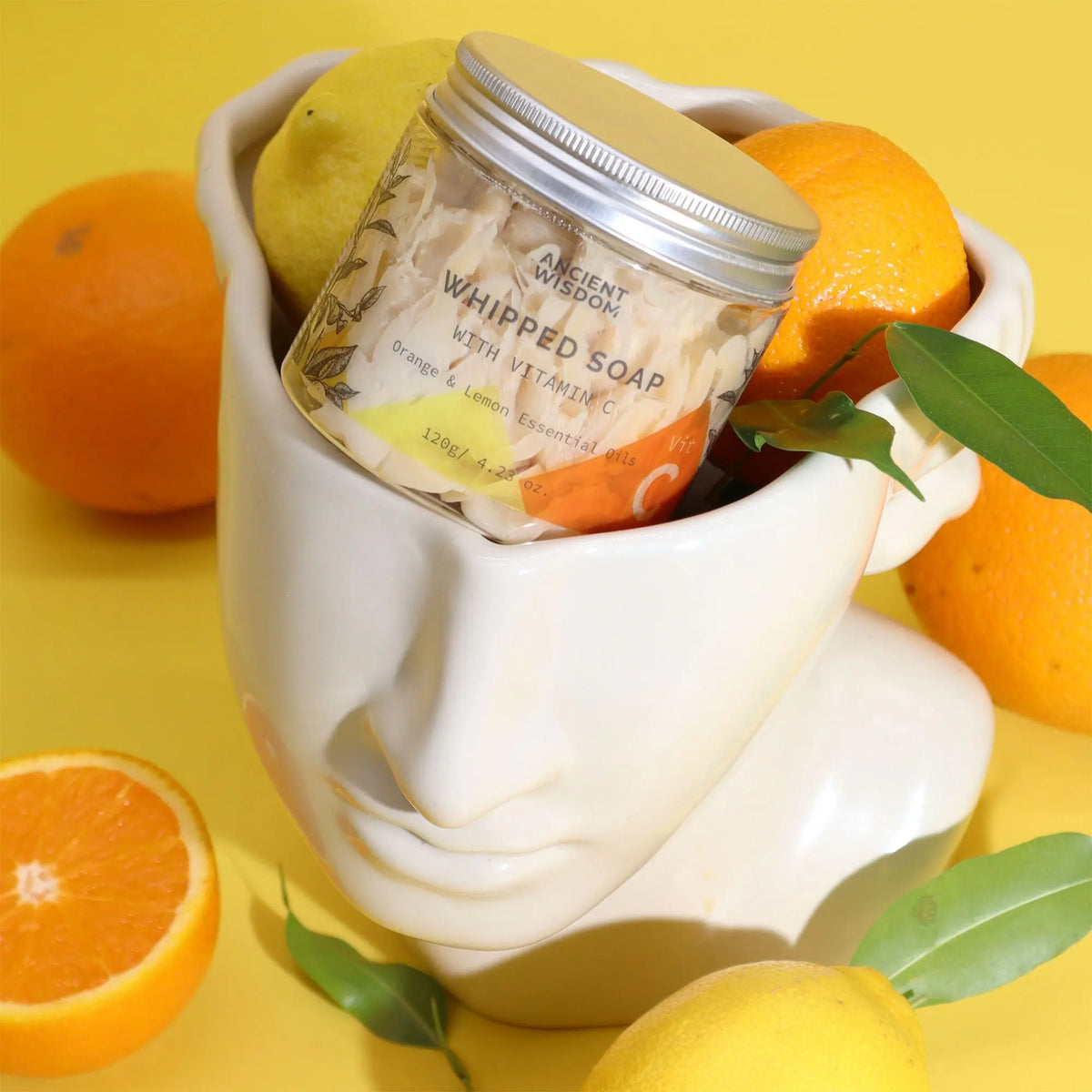 Tarro de jabón batido Vitamina C Ancient Wisdom en maceta con forma de cara, rodeado de naranjas y limones amarillos.