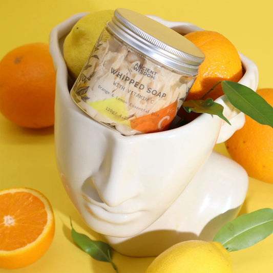 Tarro de jabón batido Vitamina C Ancient Wisdom en maceta con forma de cara, rodeado de naranjas y limones amarillos.
