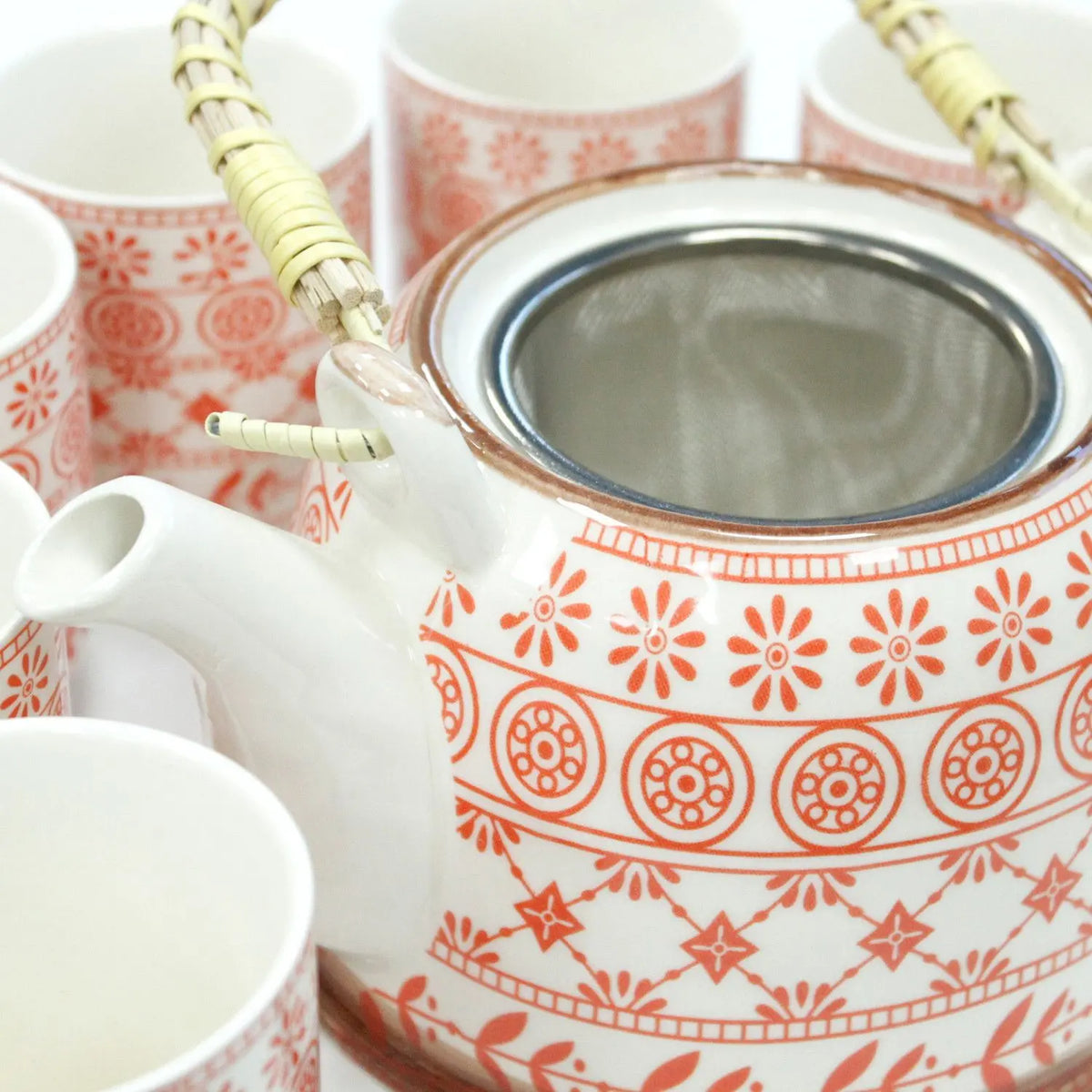 Tetera de cerámica blanca con estampado floral naranja, colador metálico y asa de mimbre, rodeada por seis tazas.
