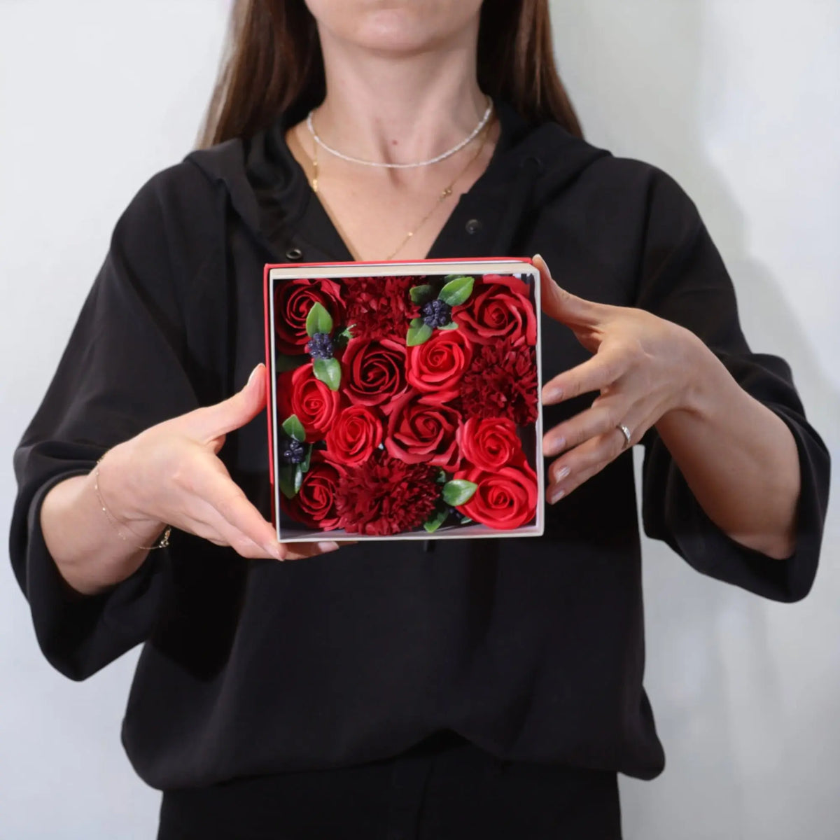 Persona sosteniendo una caja cuadrada con rosas y flores de jabón rojas, estilo elegante y detalle decorativo natural