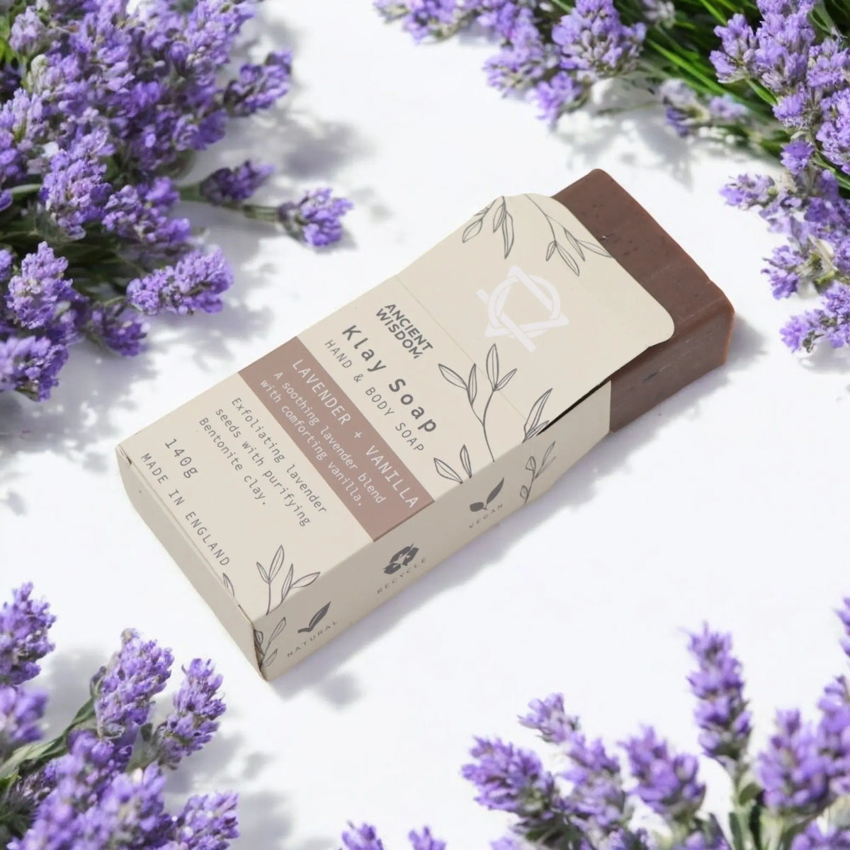 Jabón artesanal de arcilla marrón con empaque beige ilustrado, rodeado de ramilletes de lavanda sobre fondo blanco.