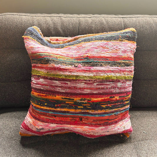 Cojín cuadrado (40x40cm) hecho de tiras de tapete reciclado con franjas horizontales en tonos azules, rojos y rosas.