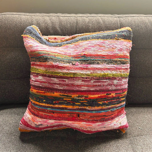 Cojín cuadrado (40x40cm) hecho de tiras de tapete reciclado con franjas horizontales en tonos azules, rojos y rosas.