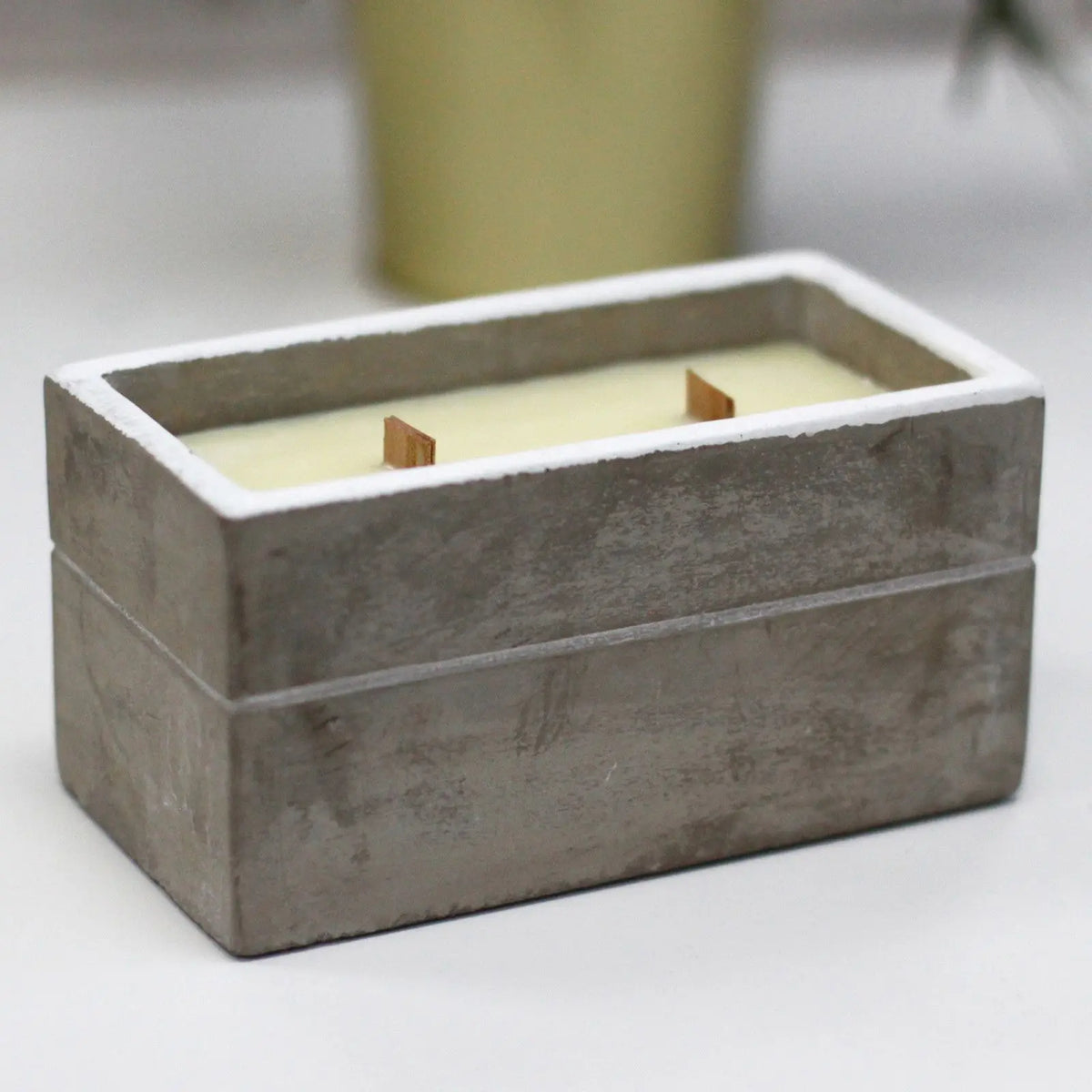 Vela rectangular en recipiente de hormigón gris con borde blanco y dos mechas de madera sobre fondo claro