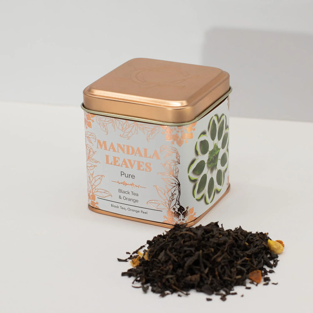 Lata cuadrada de metal color cobre con diseño floral y hojas de té negro con trozos de naranja sobre fondo blanco