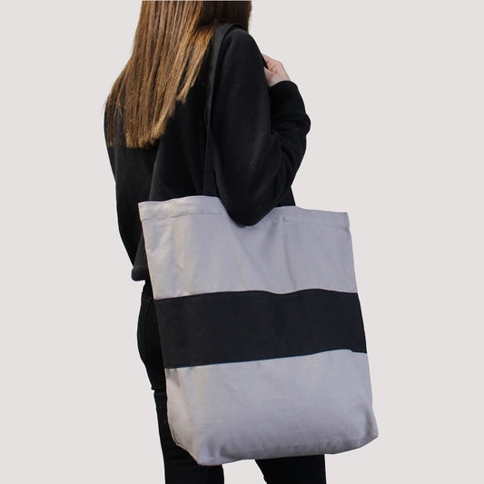 Persona cargando bolsa de algodón bicolor gris y negra, estilo tote bag, con asas largas y diseño resistente.
