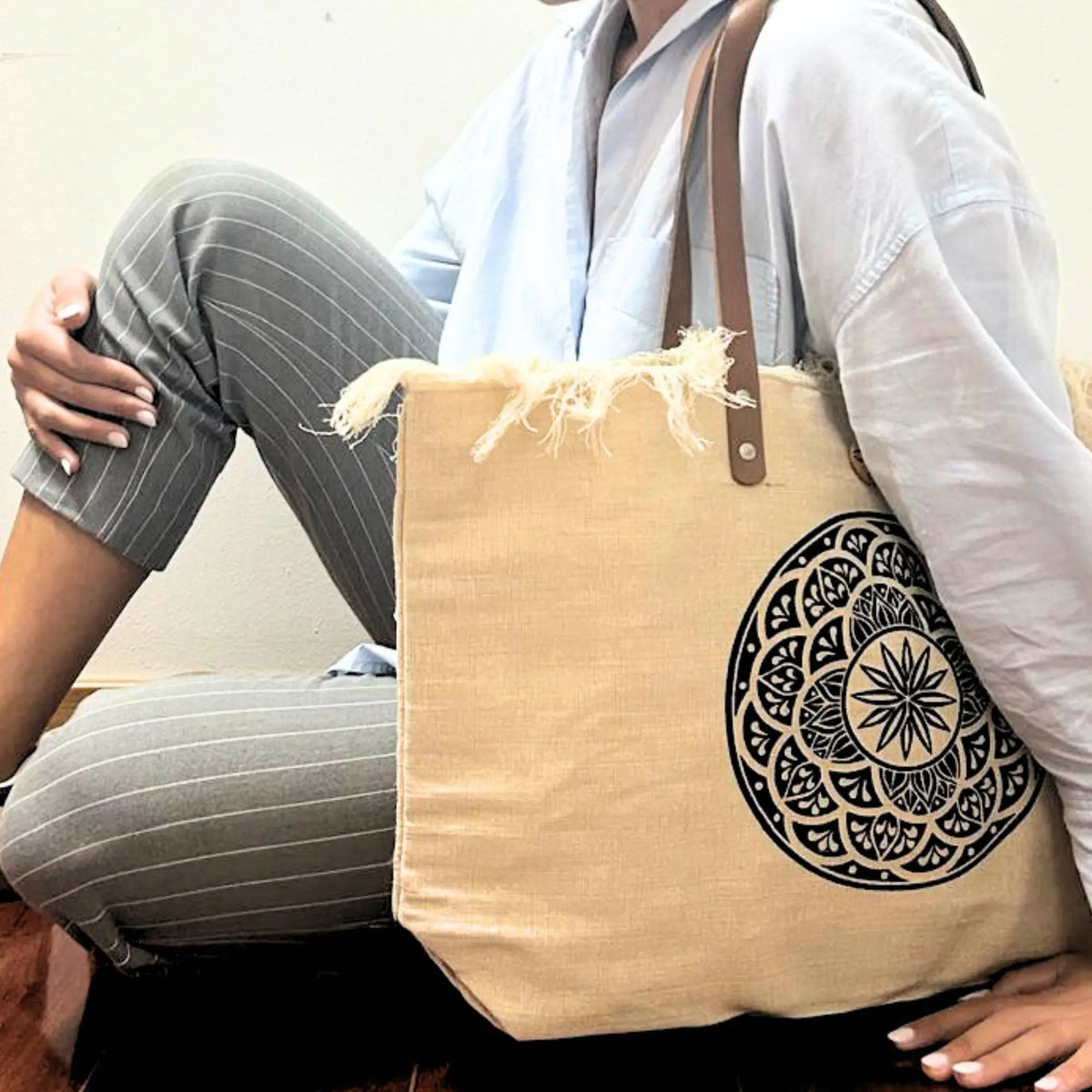 Bolso de tela natural color beige con diseño de mandala negro y asas de piel marrón, con detalle de flecos en la parte superior.