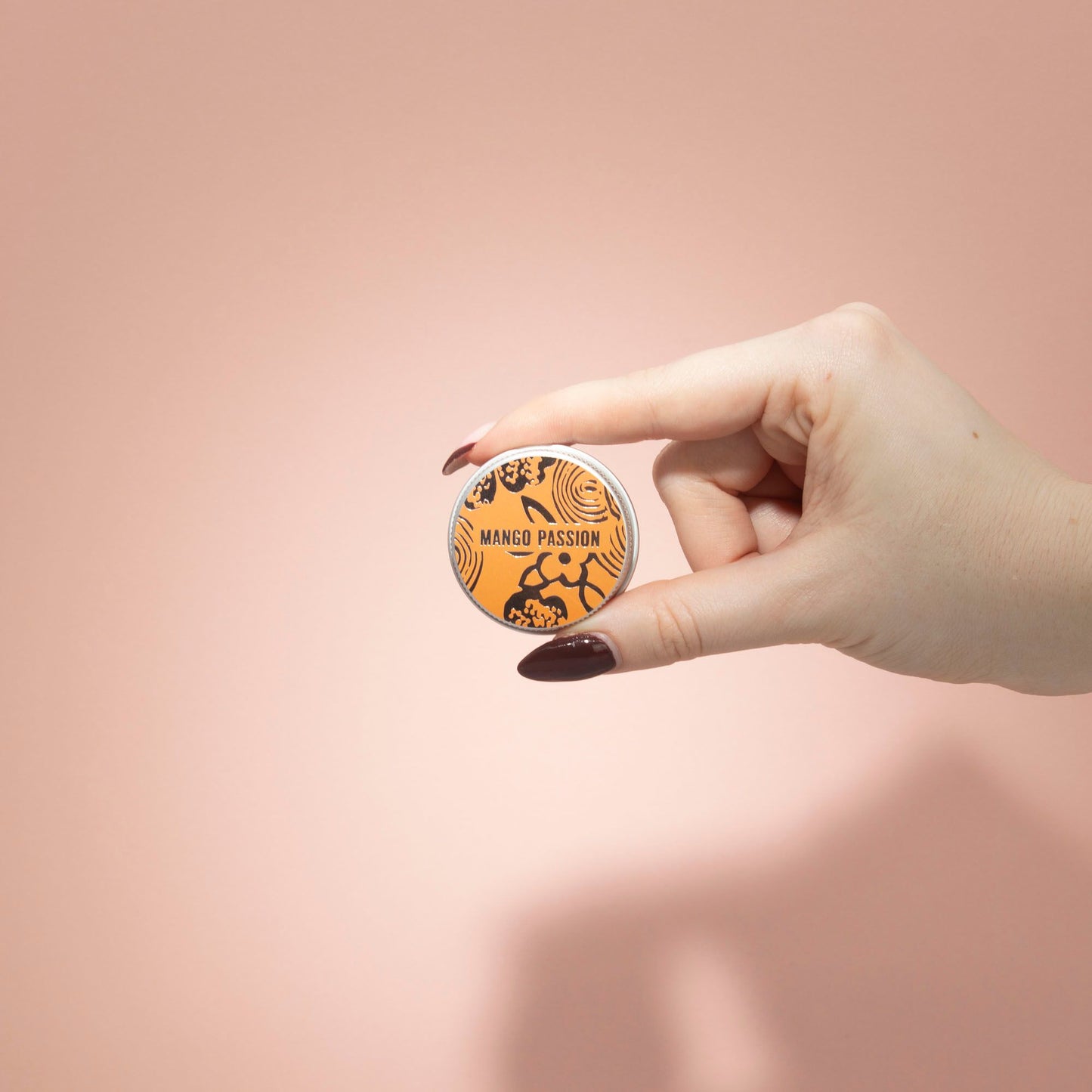 Mano sostiene pequeña lata de bálsamo labial naranja de Ancient Wisdom con diseño floral y texto "MANGO PASSION".