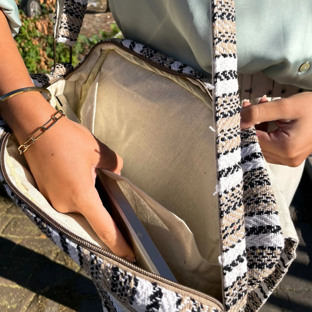 Bolso de hombro tejido en blanco, gris y beige con interior claro, donde una mano guarda un portátil fino.