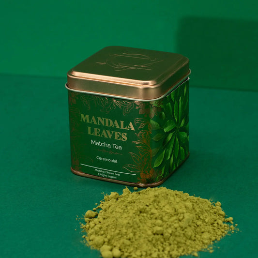 Lata cuadrada verde con detalles dorados y polvo de té matcha esparcido sobre un fondo verde uniforme.