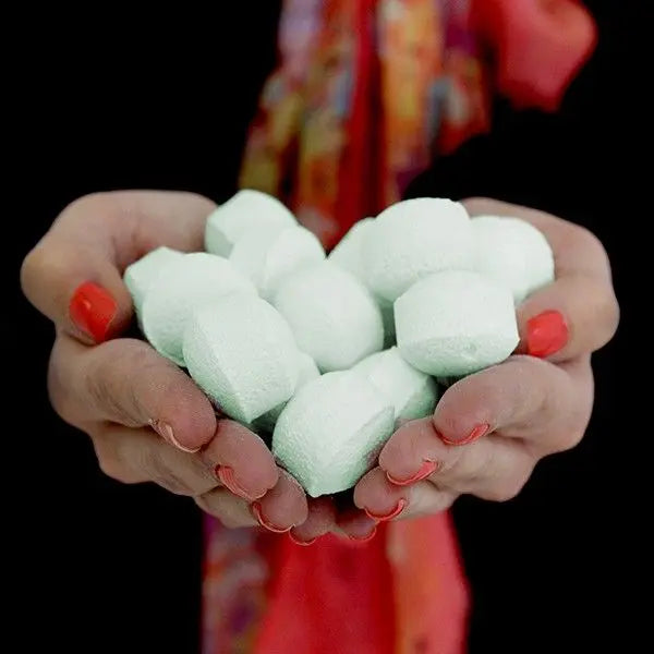 Manos sosteniendo un puñado de pequeñas bombas de baño efervescentes de color verde claro y forma circular.