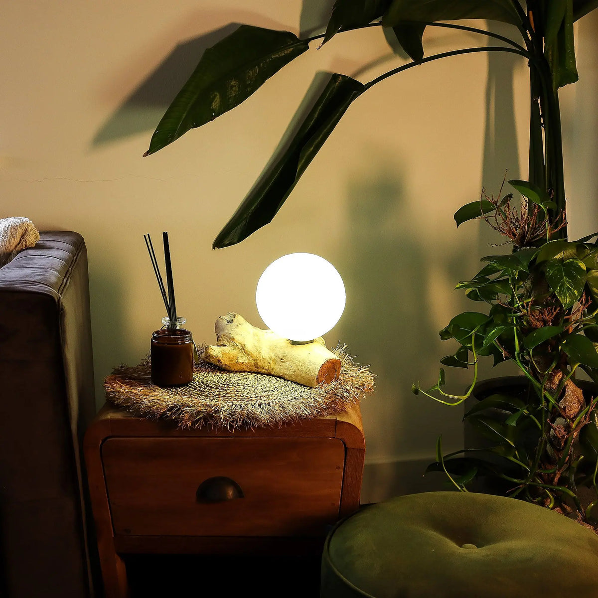 Lámpara de mesa con pantalla esférica blanca sobre una base de madera natural, encendida sobre una mesita de noche.