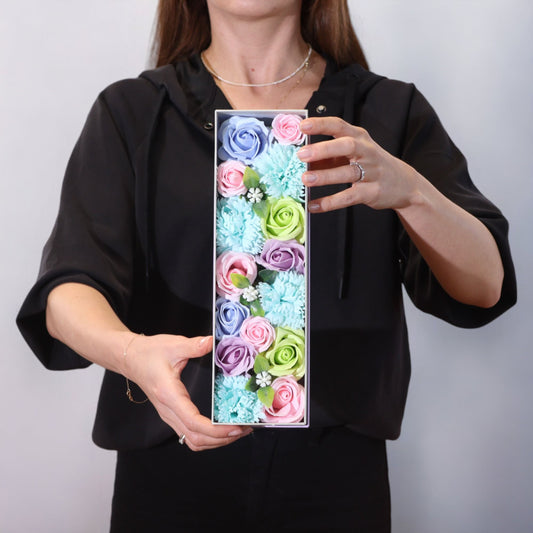 Persona sosteniendo una caja rectangular con flores de jabón multicolores en tonos pastel, azul, rosa y verde.