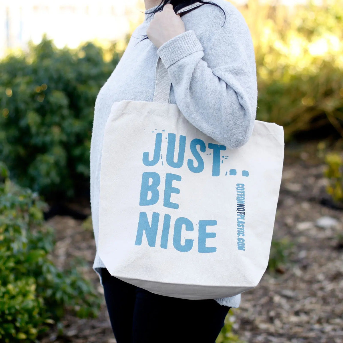 Bolsa de algodón color beige con el texto en azul "JUST BE NICE" colgada al hombro sobre un fondo de jardín.