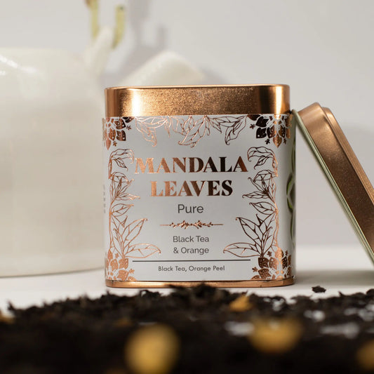 Lata metálica color bronce con detalles florales, rodeada de hojas de té negro suelto sobre un fondo blanco minimalista