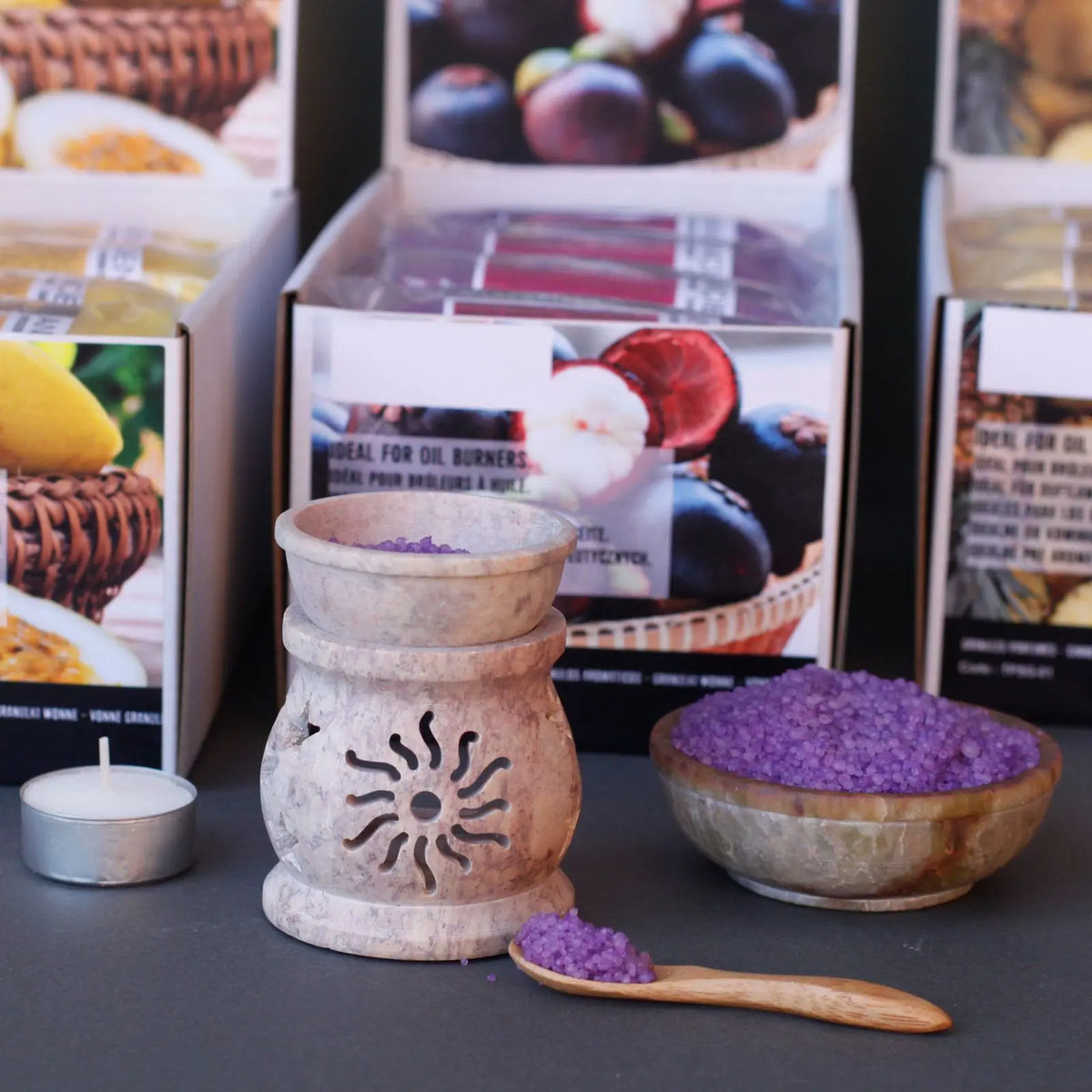 Quemador de piedra con diseño de sol, cuenco con gránulos aromáticos morados y una cuchara de madera sobre mesa gris.