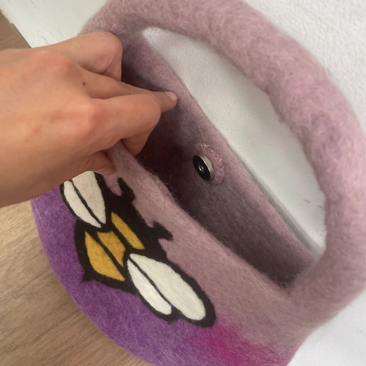 Bolso de fieltro morado con diseño de abeja, asa integrada y cierre magnético, sostenido por una mano.