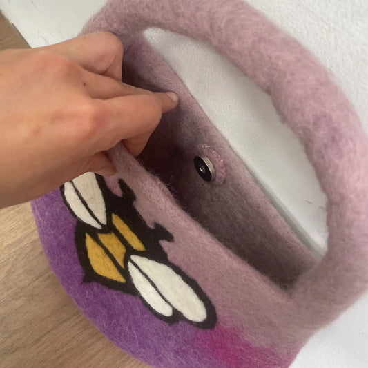 Bolso de fieltro morado con diseño de abeja, asa integrada y cierre magnético, sostenido por una mano.