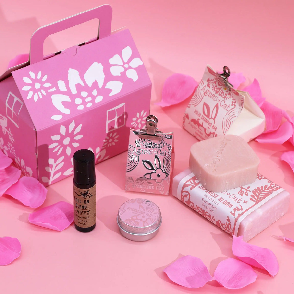 Set de regalo Agnes & Cat en caja rosa con pétalos, jabones, champú sólido, aceite roll-on y lata sobre fondo rosa.
