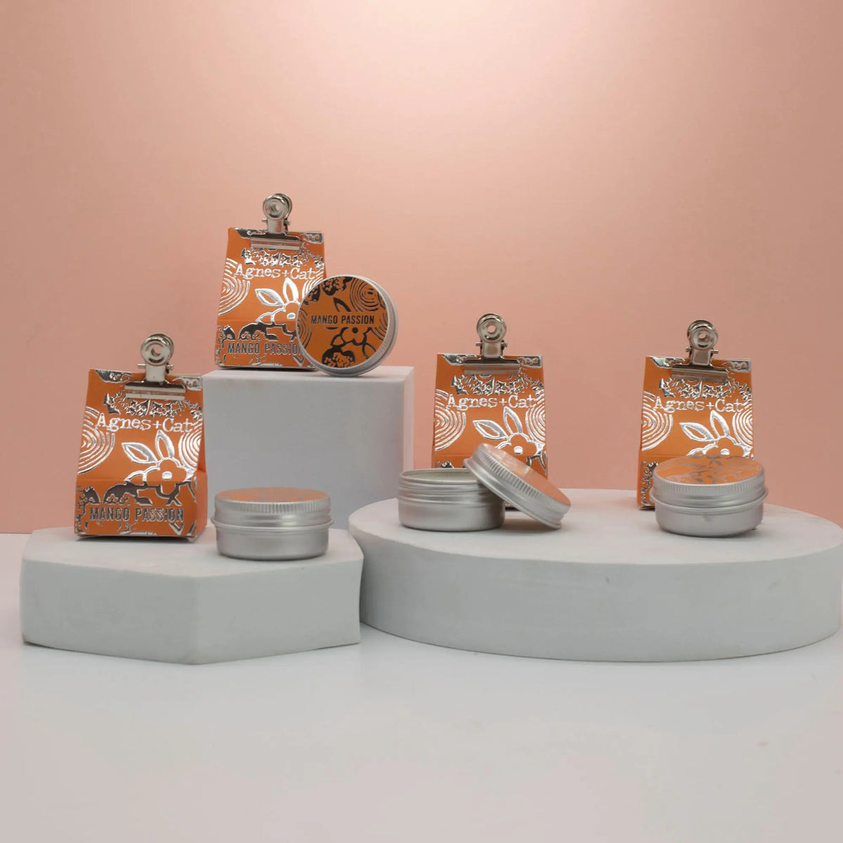Bálsamos labiales Agnes + Cat Mango Maracuyá en latas metálicas y bolsas naranjas decoradas sobre pedestales blancos.