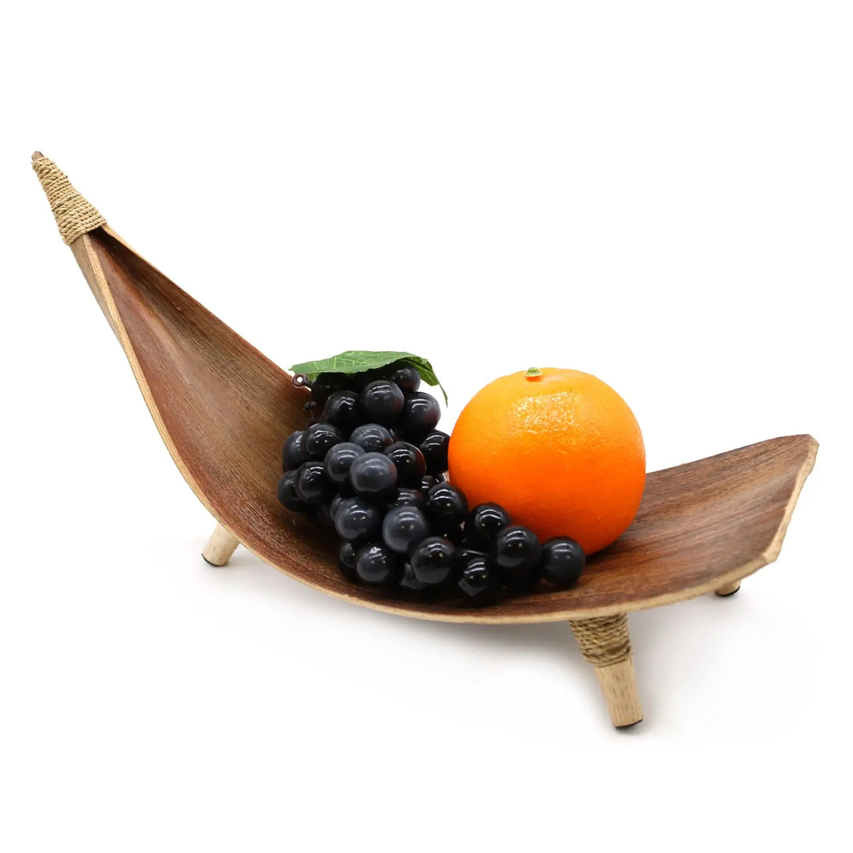 Frutero curvo de hoja de coco natural con base de tres patas, conteniendo un racimo de uvas y una naranja sobre fondo.