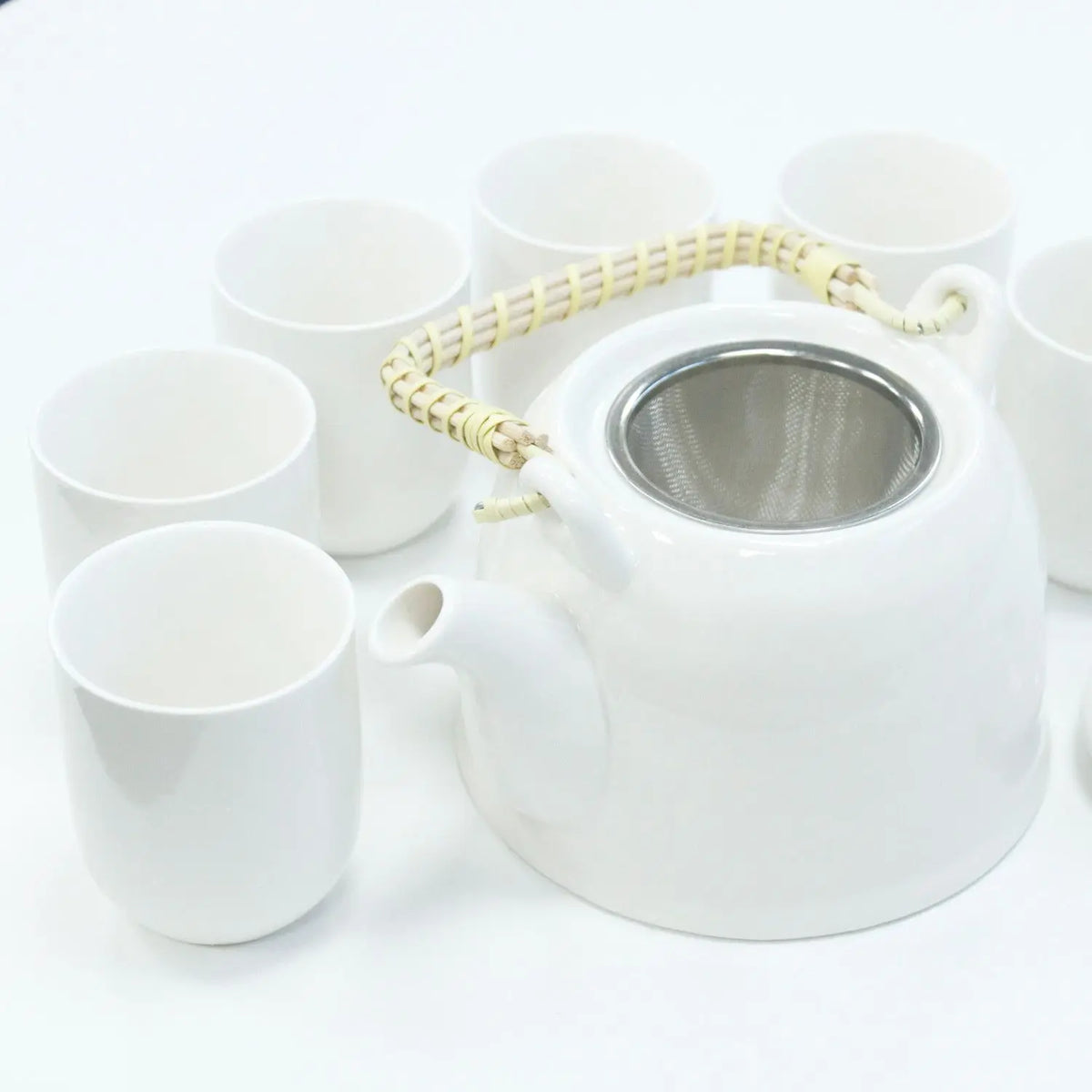 Set de té Ancient Wisdom: tetera blanca de cerámica con filtro metálico y asa de mimbre, rodeada de tazas a juego.