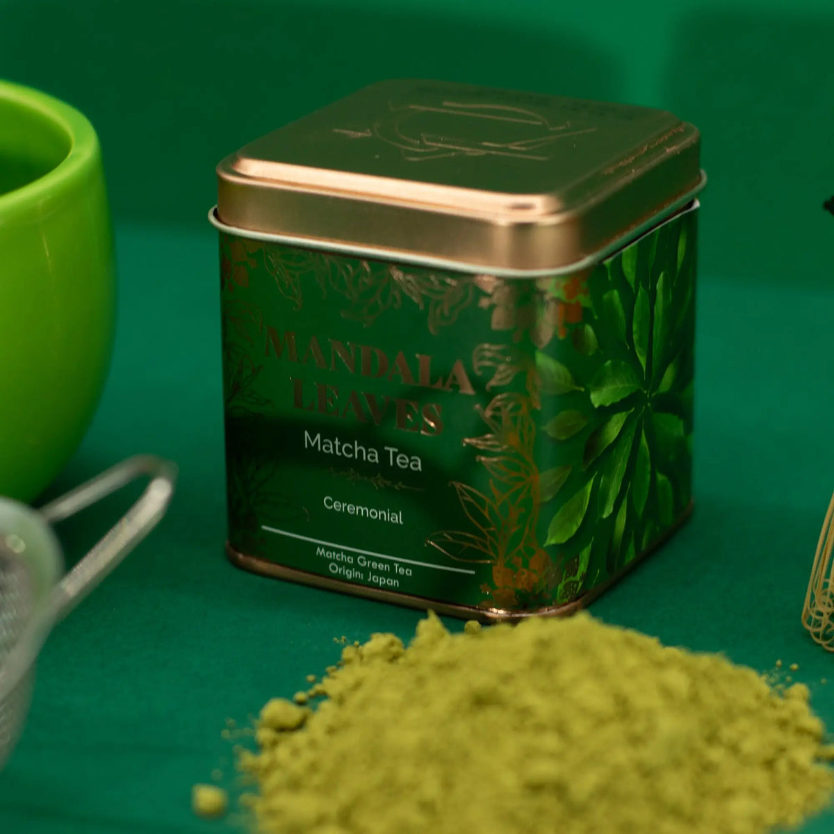 Lata cuadrada verde de té matcha ceremonial con detalles dorados, junto a polvo de matcha sobre fondo verde.