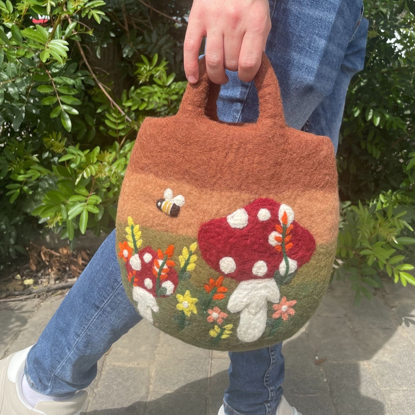 Bolsa de fieltro marrón con diseño de setas y flores en relieve, sostenida por una persona al aire libre.