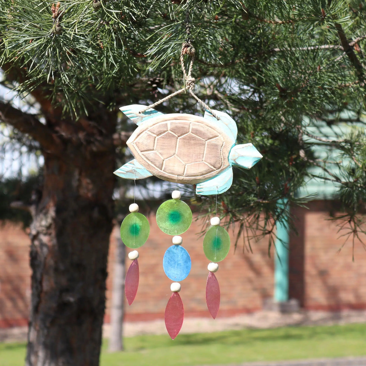 Carillón de madera con forma de tortuga color turquesa y colgantes de cuentas circulares y gotas de colores en un árbol.